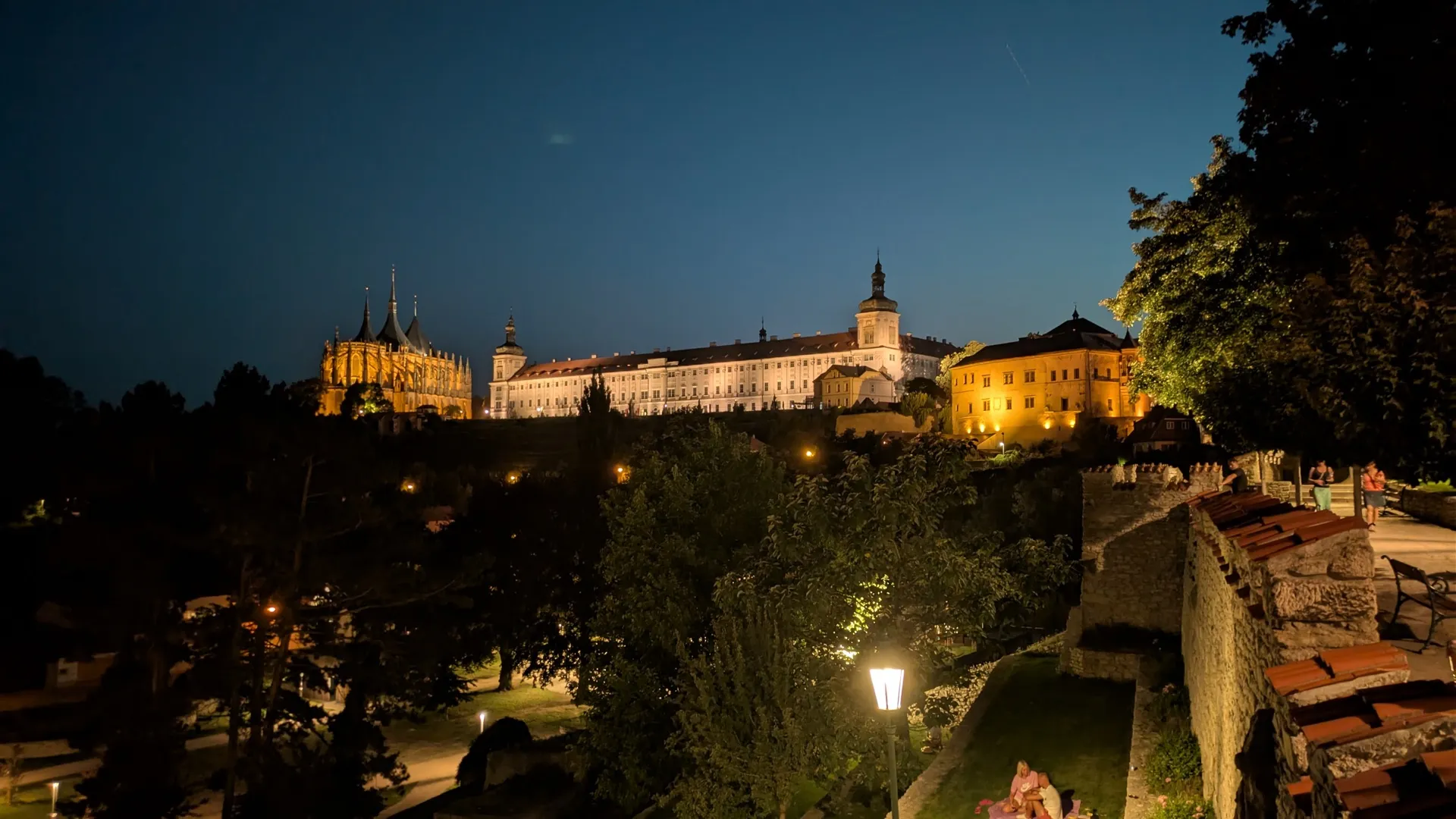 Noční panorama Kutné Hory od hradeb: vlevo nasvícený chrám svaté Barbory, uprostřed dlouhá budova Jezuitské koleje, vpravo hradby s lidmi a pouliční lampa, nad městem tmavnoucí modrá obloha po západu slunce